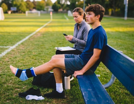 A young boy rests on a bench with an injured ankle wrapped in a bandage during football practice.